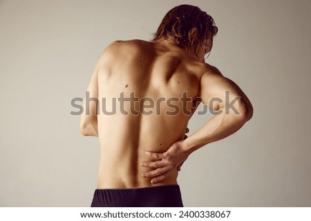 Lower back pains. Rear view of shirtless young man suffering from back pains against grey studio background. Concept of men's beauty, health, body care, sportive lifestyle, medicine Royalty-Free Stock Photo #2400338067