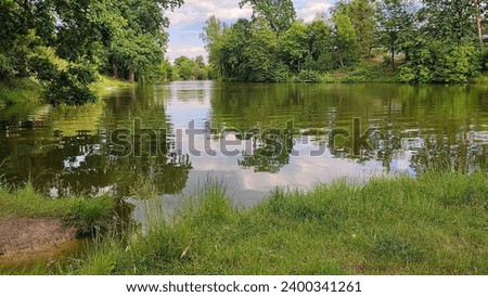 beautiful panorama of a pond with water in a green forest against the backdrop of trees without people and part of the shore with green grass in the summer afternoon Royalty-Free Stock Photo #2400341261