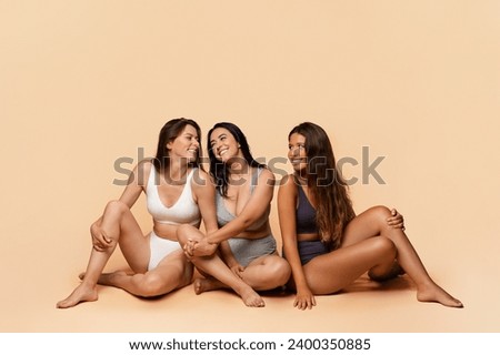 Three glad diverse millennial women in undergarments sit on floor, enjoy body positivity, beauty care, have fun, isolated on light studio background, full length. Friendship, confidence Royalty-Free Stock Photo #2400350885