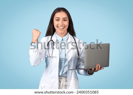 Happy woman doctor holding computer makes a triumphant success gesture, possibly celebrating medical advancements or achievements, blue background Royalty-Free Stock Photo #2400351085