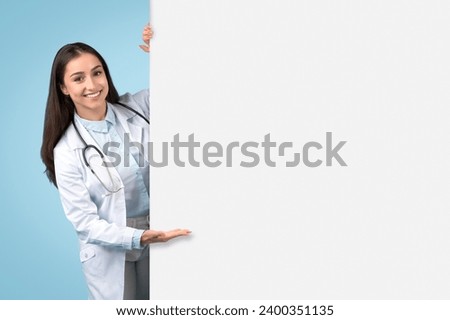 Cheerful female doctor in white coat showcases a blank space for medical advertising or informational content, presenting placard board on blue background Royalty-Free Stock Photo #2400351135