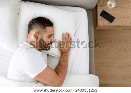 Rest and relaxation. Man enjoys healthy sleep routine, lying in cozy white bed in modern bedroom indoor, smartphone lying near table. Oversleeping concept. Copy space for text