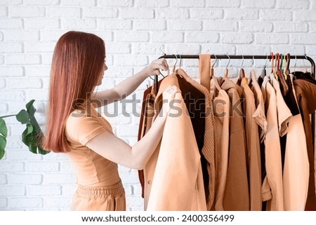 Color of the year 2024: Peach Fuzz. beautiful young woman in beige coat standing in front of hanger rack and choosing outfit dressing. Selection of a wardrobe, stylist, shopping. Clothes designing Royalty-Free Stock Photo #2400356499