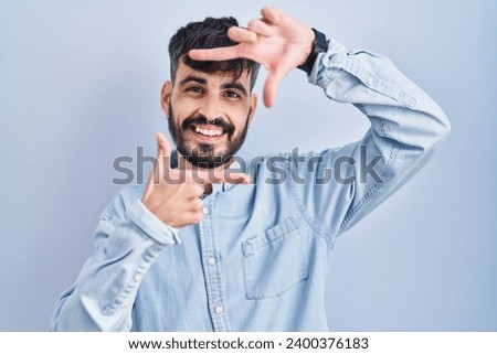 Young hispanic man with beard standing over blue background smiling making frame with hands and fingers with happy face. creativity and photography concept. 