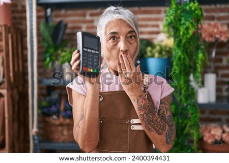 Middle age woman with tattoos working at florist shop holding dataphone covering mouth with hand, shocked and afraid for mistake. surprised expression 