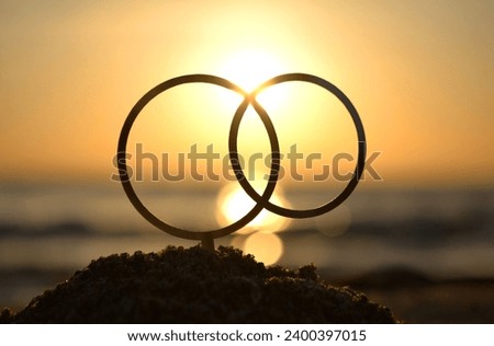 Black outlines of wedding rings at dawn and sunset on seashore. Stick contours in shape of wedding rings in sand on backdrop of setting and rising sun. Concept wedding love infatuation Valentine's Day Royalty-Free Stock Photo #2400397015
