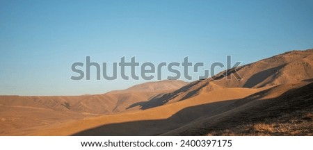 Sunset light casts a warm glow on the smooth, undulating contours of a grass-covered highland, with shadowy mountain ridges in the background under a soft sky Royalty-Free Stock Photo #2400397175
