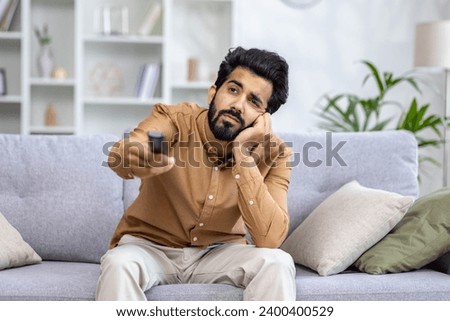 A young Indian man sits and rests tiredly at home on the sofa, uses the TV remote control, lazily rests his head on his hand, looks bored into the window.