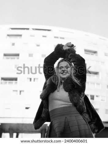 Smiling female model posing with white building behind in a cold winter day. Her arms are up and the image is monochromatic, made with an old film camera.