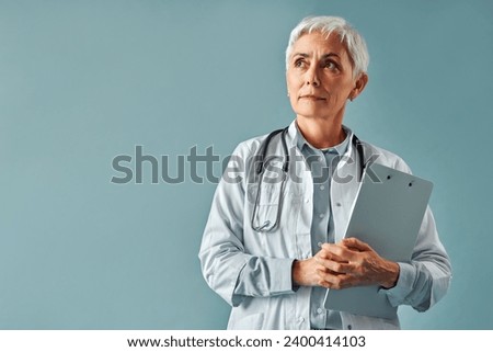 A mature female doctor in a white coat stands on a white background, holds a tablet in her hands and looks seriously thoughtfully to the side at the space for text, advertising.                       