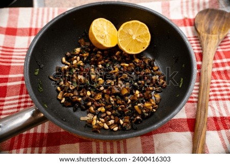 Pan of fried bacon, onions, mushrooms and pine nuts garnished with sage, parsley and two lemon halves on a red checkered background