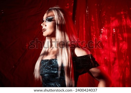 Side view portrait of extravagant drag queen standing on stage in spotlight against red curtains, copy space Royalty-Free Stock Photo #2400424037