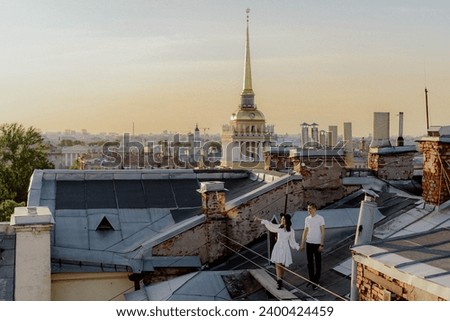A man and woman walking, hugging and kissing on a roof. Rooftop in Saint-Petersburg, Russia. View at Admiralty. Young couple romantic date in a city at sunset. Royalty-Free Stock Photo #2400424459