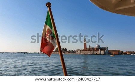 Waving Italian flag with scenic view church on the Island of San Giorgio Maggiore, Venice, Veneto, Northern Italy, Europe. Perspective from ferry boat trip. Patriotic banner moving to the wind