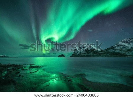 Aurora borealis over the sea, snowy mountains at starry winter night. Northern Lights in Lofoten islands, Norway. Sky with polar lights. Landscape with aurora, rocky beach, sky, reflection in water Royalty-Free Stock Photo #2400433805