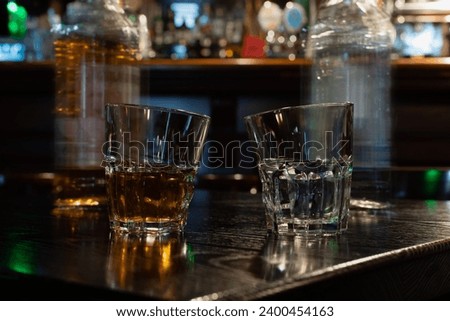 Two curved original unusual cut glasses with gold and transparent spirit drinks on dark wooden table against blurred irish pub reflections