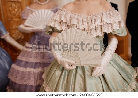 Closeup of ladies wearing big ballgowns holding fans while waiting for partners during ball in palace, copy space Royalty-Free Stock Photo #2400466563