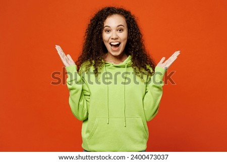 Young surprised shocked amazed overjoyed woman of African American ethnicity she wears green hoody casual clothes look camera spread hands isolated on plain red orange background. Lifestyle concept