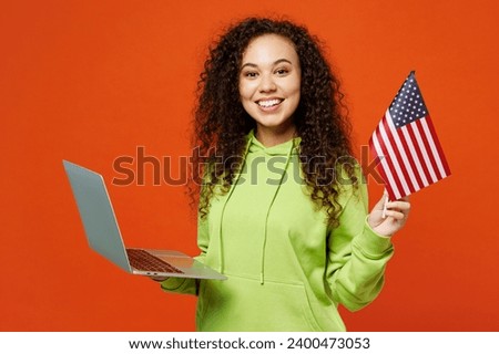 Young smiling IT woman of African American ethnicity she wear green hoody casual clothes hold American flag use work on laptop pc computer isolated on plain red orange background. Lifestyle concept