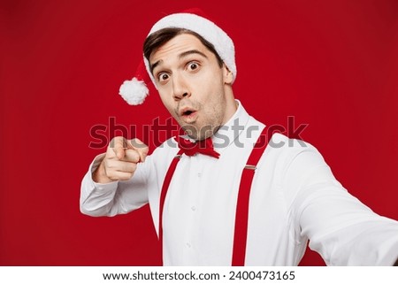 Close up merry young man wear white shirt Santa hat posing doing selfie shot pov mobile cell phone point finger camera on you isolated on plain red background. Happy New Year Christmas holiday concept