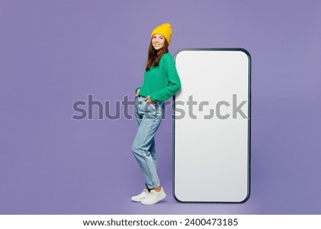 Full body young happy woman she wear green sweater yellow hat casual clothes stand near big huge blank screen mobile cell phone smartphone with area isolated on plain pastel light purple background