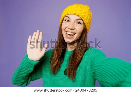Close up young woman she wear green sweater yellow hat casual clothes doing selfie shot pov on mobile cell phone waving hand isolated on plain pastel light purple background studio. Lifestyle concept