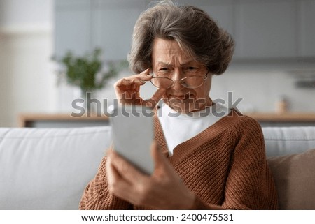 Senior european woman squinting eyes looking at phone screen, wearing eyeglasses sitting on couch at home. Bad vision health problems Royalty-Free Stock Photo #2400475531