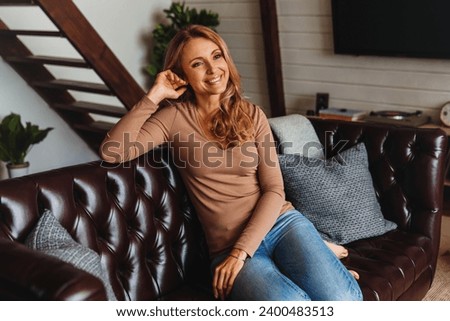 Happy adult woman relaxing on her couch at home in living room. Middle-aged mature caucasian woman taking rest, spending me-time in apartment. Self care Royalty-Free Stock Photo #2400483513
