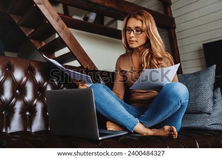 Middle aged woman working at home sitting on sofa in the living room with laptop and checking documents, bills, debts, financial papers, mortgage, bank loans remotely online Royalty-Free Stock Photo #2400484327