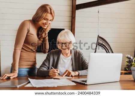 Beauty middle aged lady teaching her mom to use computer, pay domestic bills together online, filling in health insurance, loan, receiving pension on bank account Royalty-Free Stock Photo #2400484339