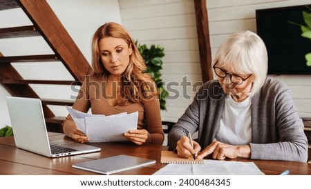 Senior woman with help of her daughter making bills at home, paying loan, debt, filling in medical insurance, mortgage documents, health insurance Royalty-Free Stock Photo #2400484345