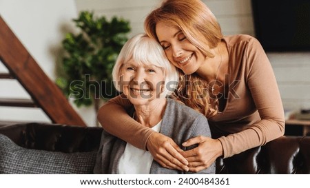 Happy daughter embracing from behind elderly mother at living room. Love and care. Family time and bonding concept. Assistance and support of elder generation Royalty-Free Stock Photo #2400484361