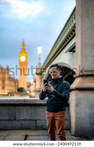 Happy young Latin man tourist in casual clothes standing alone against ancient cathedral in London near tower bridge and Big Ben against clock tower and using smartphone