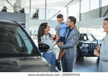 Young happy family enjoying while buying a new car in a showroom. Saleswoman at car dealership center helping family to choose new family vehicle. Family in a car dealership  Royalty-Free Stock Photo #2400494761