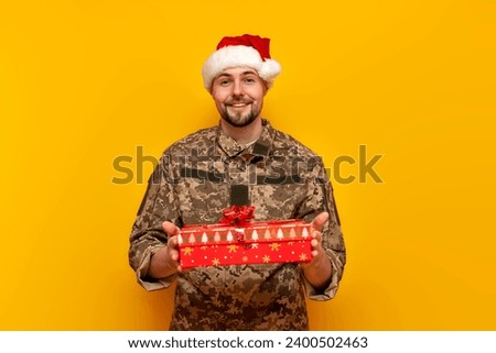 Ukrainian soldier in pixel uniform and santa claus hat gives a gift box on a yellow isolated background, military man of the ukrainian army with a gift and donation congratulates for christmas