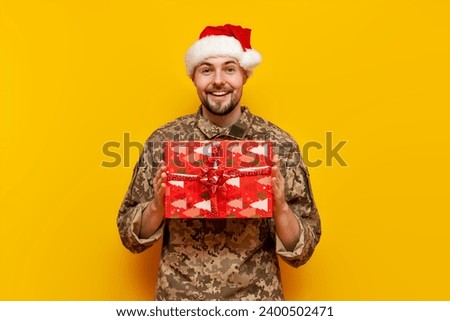 Ukrainian soldier in pixelated uniform and santa claus hat holding gift box on yellow isolated background, ukrainian army military man with gift and donation for christmas