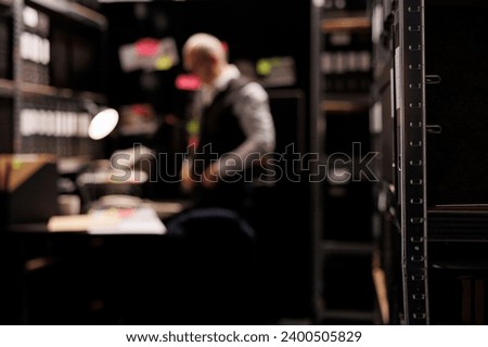 Selectiv focus of metallic shelves full with police documents, in background private detective working overhours at criminal investigations in arhive room. Elderly inspector analyzing crime evidence