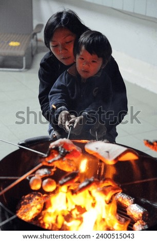 Exterior photo view of a mom mum mother and her son child kid boy cooking roasting grilling food meat together on a bbq barbecue by night with friends or family arounf during gathering happy moment
