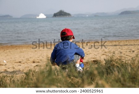 Exterior photo view of a young kid child children male active  boy playing a the beach alone by himself on the sand with his plastic toys near the sea ocean view from the back crouching
