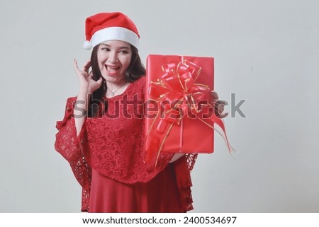 Happy mature asian woman celebrating Christmas, wearing red dress and Santa hat, while carrying gift box. on a white background.