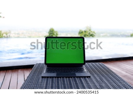 tablet computer with blank green screen on rattan woven table with blurred swimming pool background.