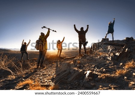 Big group of young hikers with backpacks are standing with open arms in winner poses at sunset mountain
