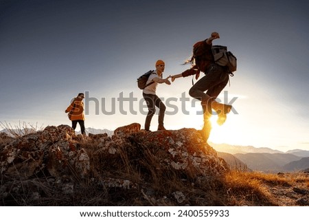 Group of young hikers with backpacks walks with backpacks and helps each other in climbing in sunset mountains Royalty-Free Stock Photo #2400559933