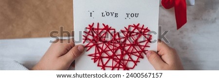 Little kid preparing diy handmade cute post card for Mother's Day with a message I love you mom. Red hear embroidering, printing. Concept of simple presents, family values, hobby, leisure activity