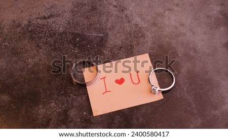 Top down view of pink adhesive note with handwritten "I love you", with a diamond ring and a plain silver ring, placed on vintage stone table surface