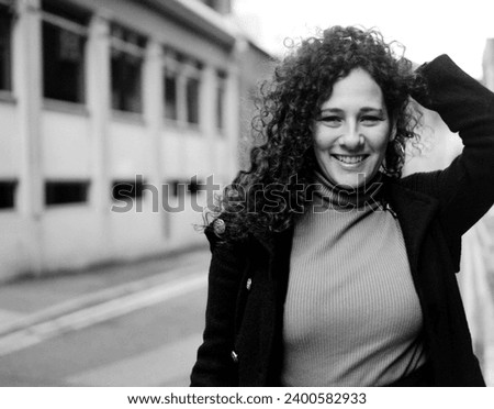 Smiling curly woman in a London street looking at camera in monochrome portrait. It is winter and she wearing coat and sweater. Image made with analog film camera.