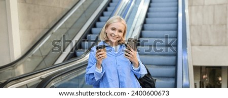 Image of stylish young woman in blue shirt, holding takeaway cup of coffee and mobile phone, standing near escalator and waiting for you, smiling at camera.