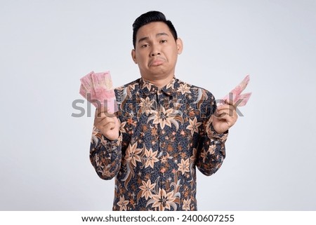Confused handsome young Asian man wearing batik shirt holding money cash isolated on white background. Profit and wealth concept