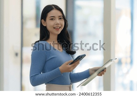 Young beautiful Asian business woman holding a folder and a smart phone while standing at the workplace.