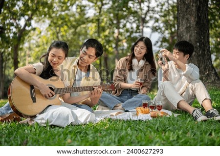 A lovely young Asian couple is playing an acoustic guitar together while enjoying a picnic with their friends in a green park on summer. Friendship concept